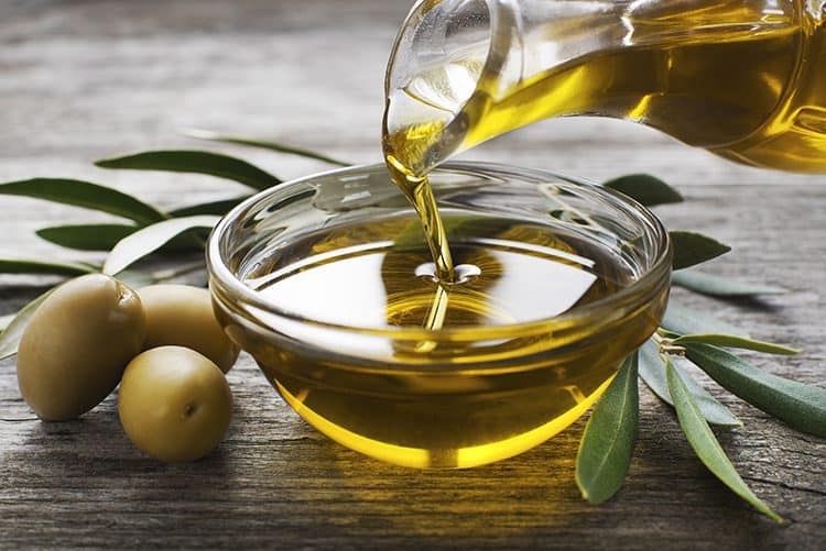 A bottle of olive oil pouring into a bowl surrounded by olives