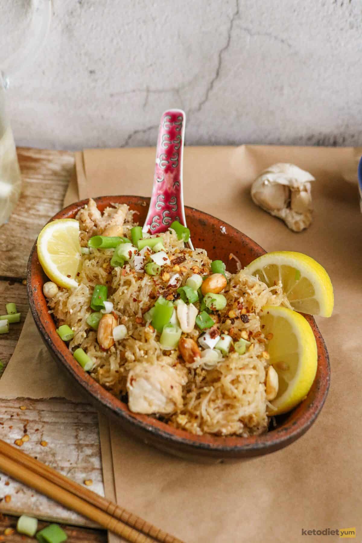 Bowl of keto chicken pad thai with a spoon and lemon wedges on a table