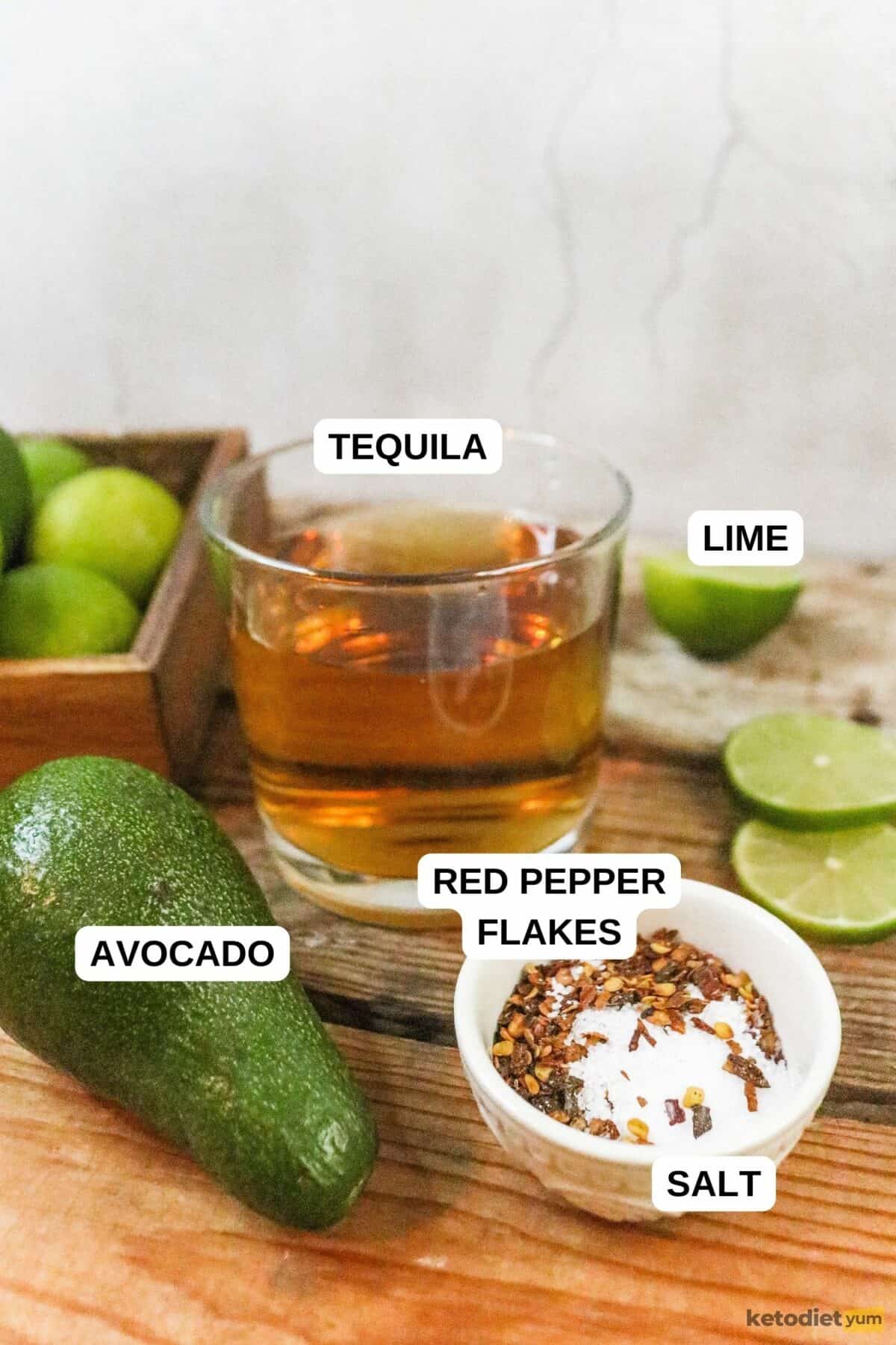 Avocado, sliced limes, whole limes, glass of tequila and bowl of red pepper flakes and salt on a table