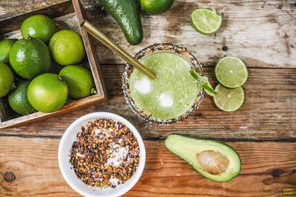 Glass with metal straw, whole limes, lime slices, half an avocado, and a bowl with red pepper salt flakes on a table