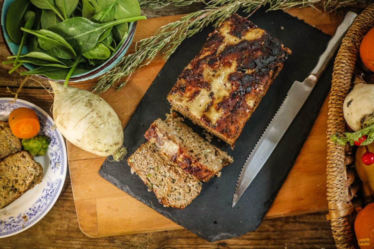 Meatloaf on a cutting board with a bowl of spinach and fresh vegetables arranged around it
