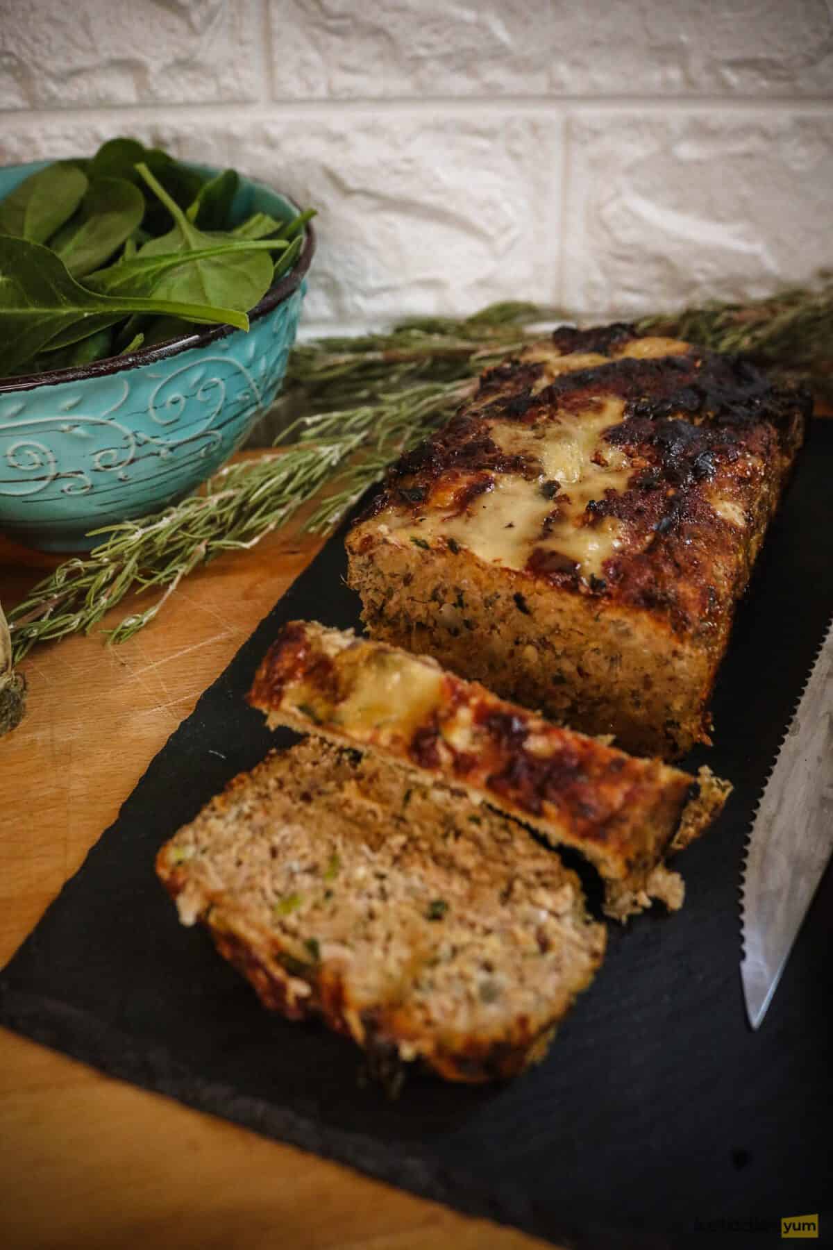 Sliced meatloaf on a serving plate with a bowl of spinach and rosemary sprigs in the background