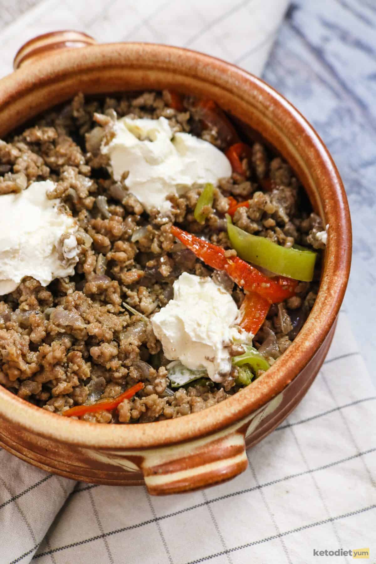 Brown casserole dish filled with ground beef, red peppers, green peppers and dollops of cream cheese on a tablecloth