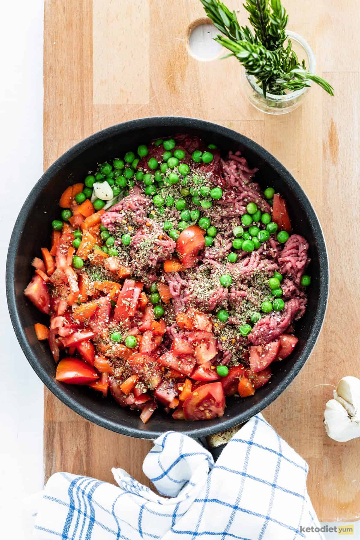 Frying pan filled with ground lamb, peas, tomatoes, carrots, herbs, and spices on top of a wooden chopping board