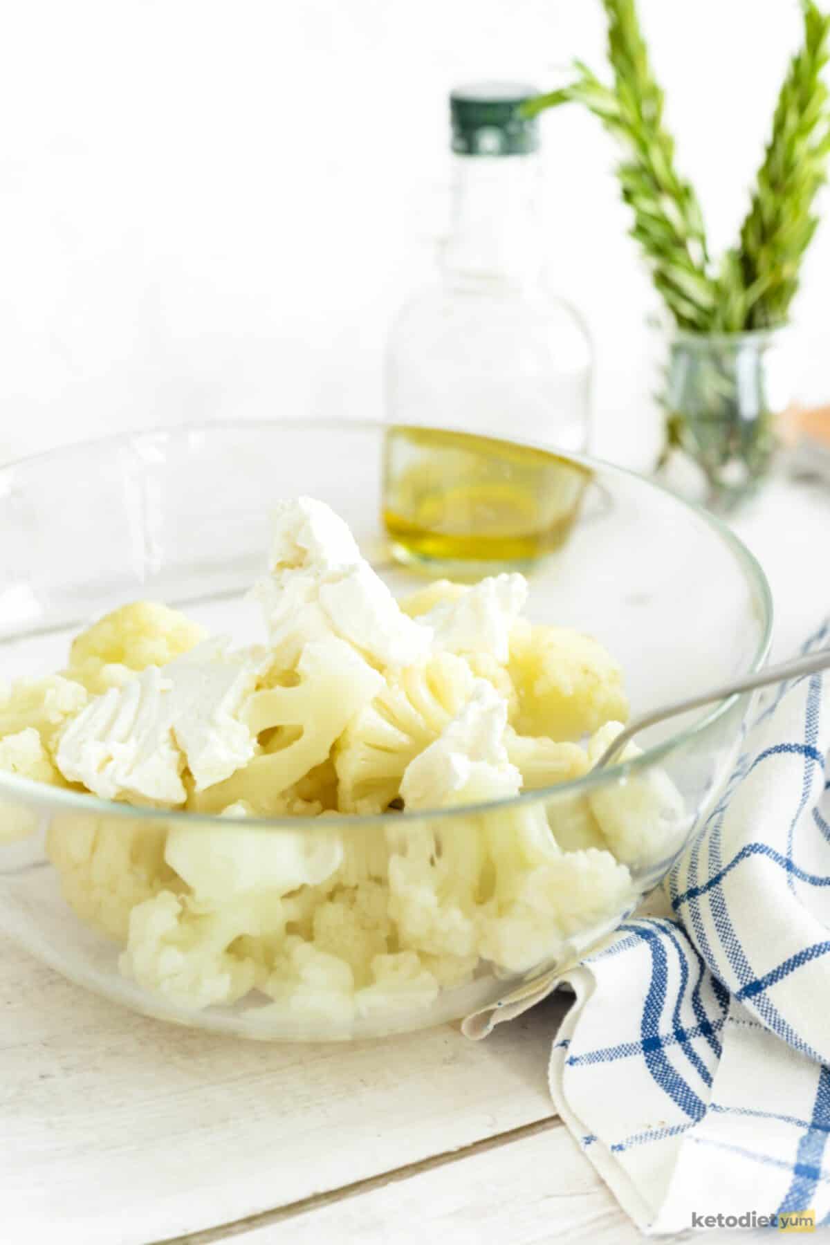 Glass bowl of cauliflower florets with a spoon inside and a kitchen towel underneath with a bottle of olive oil and jar of rosemary sprigs in the background