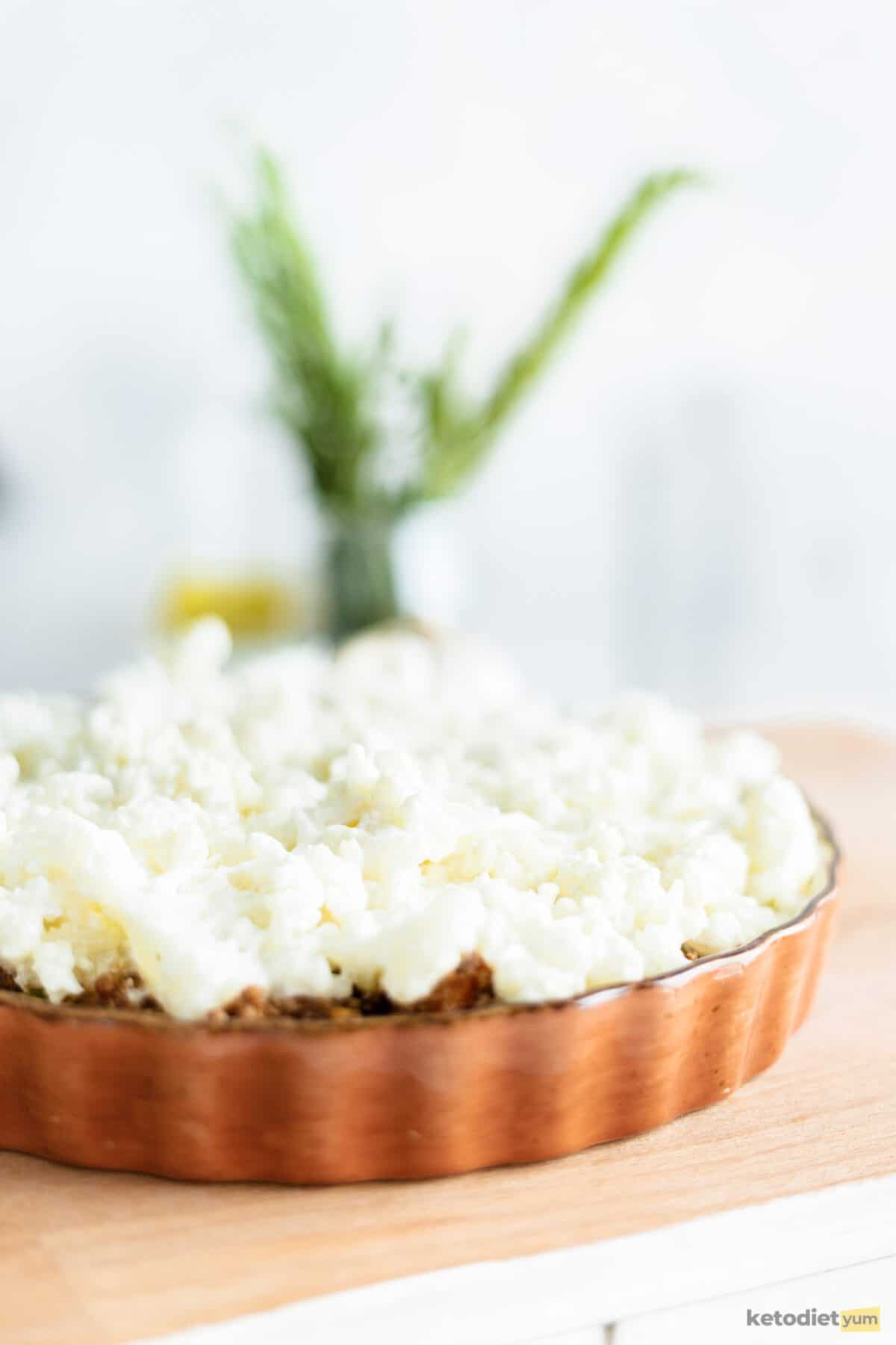 Baking dish topped with cauliflower sitting on a wooden board with a jar of fresh rosemary in the background