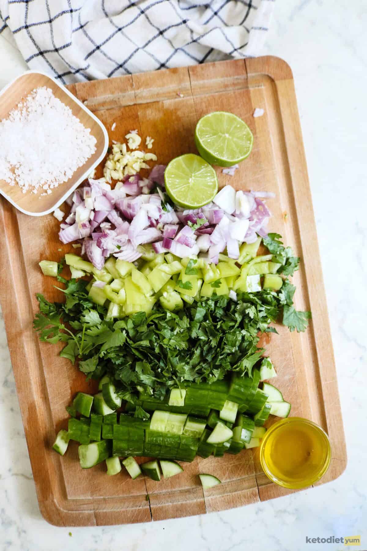 Chopping board with diced red onion, green bell pepper, cilantro, cucumber, garlic, lime, olive oil and flaky salt on a table with a kitchen cloth