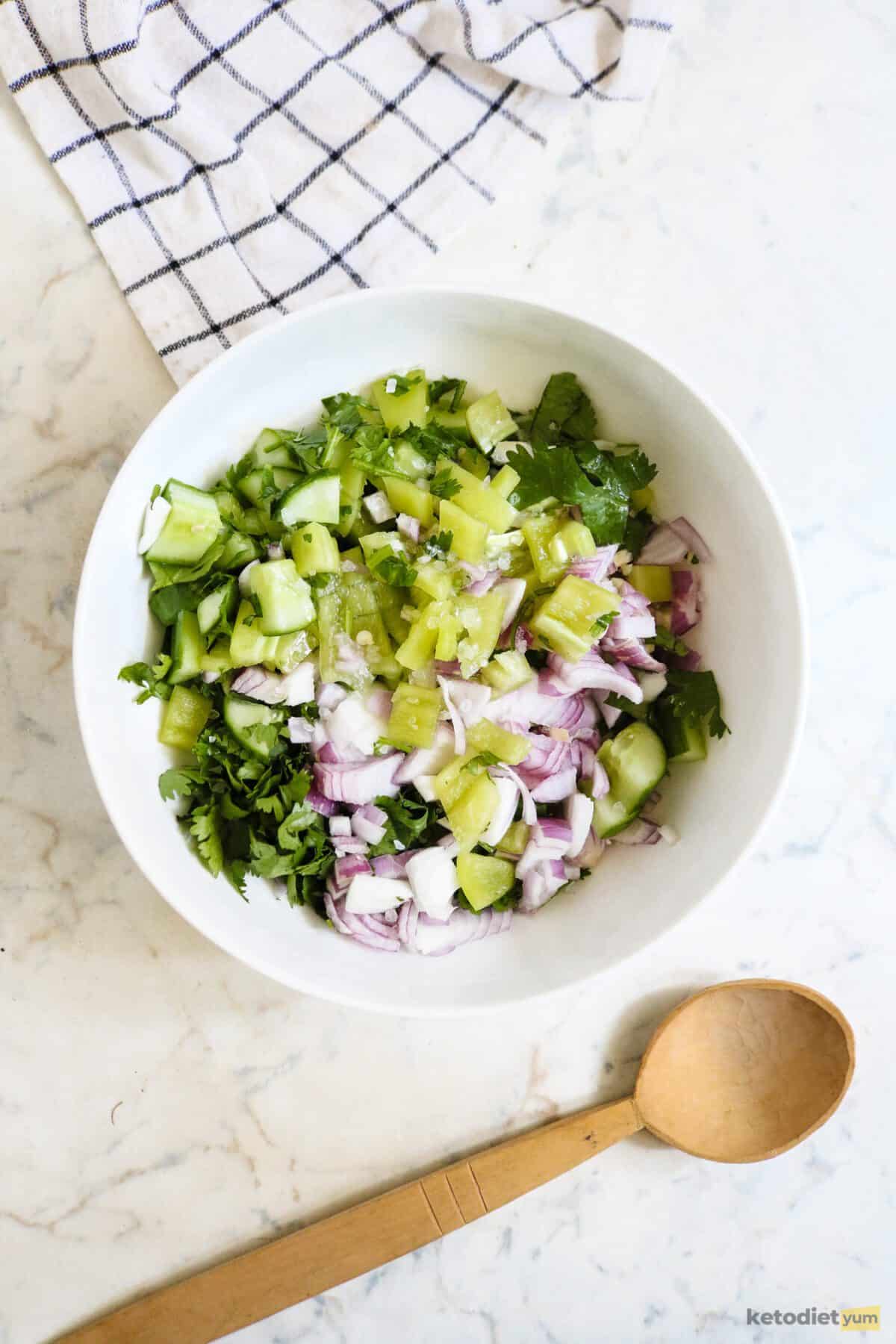 White bowl filled with diced cucumber, red onion, green pepper and jalapeno with a wooden spoon and kitchen towel