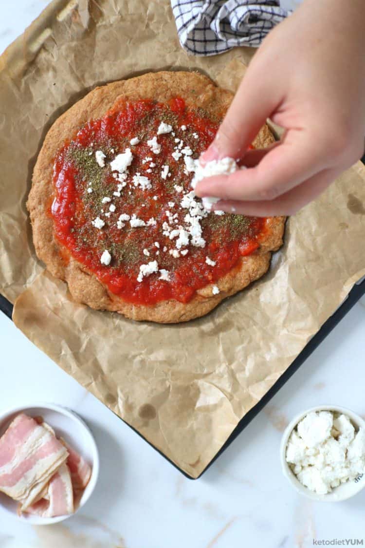 Topping the almond flour pizza crust with feta cheese and bacon