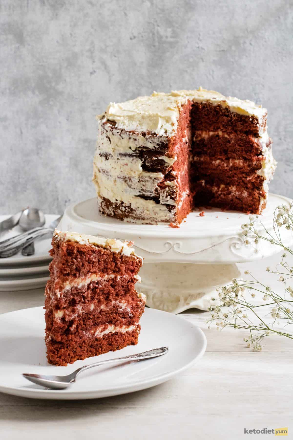 A slice of keto red velvet cake with cream cheese frosting with the full cake in the background