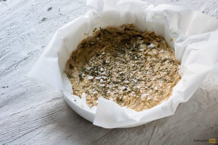 Focaccia bread dough transferred to a lined baking pan and topped with rosemary and sea salt flakes