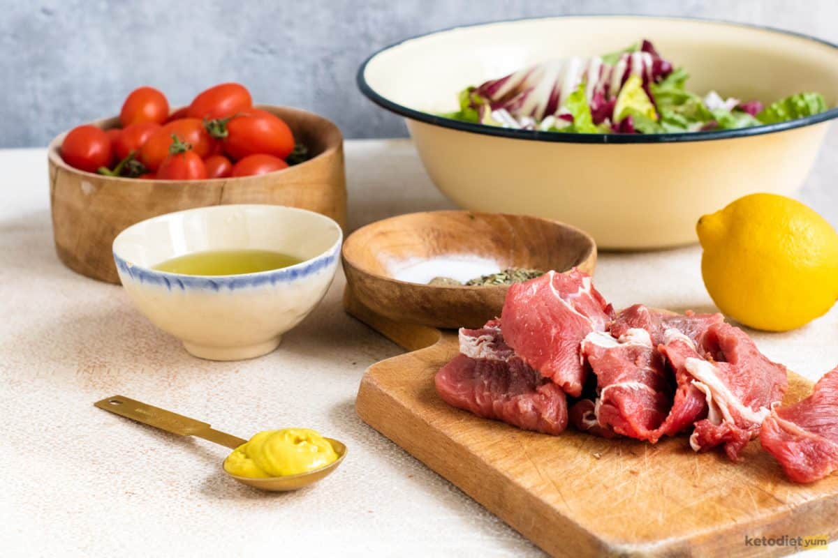 Keto steak salad ingredients arranged on a table including cherry tomatoes, sirloin steak, mixed greens, olive oil, lemon, Dijon mustard and seasonings
