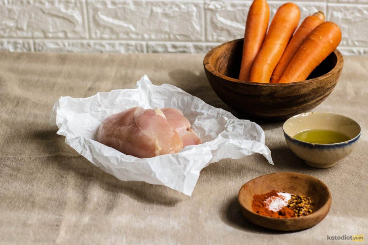 Ingredients on a table including chicken breasts, carrots, seasonings and olive oil