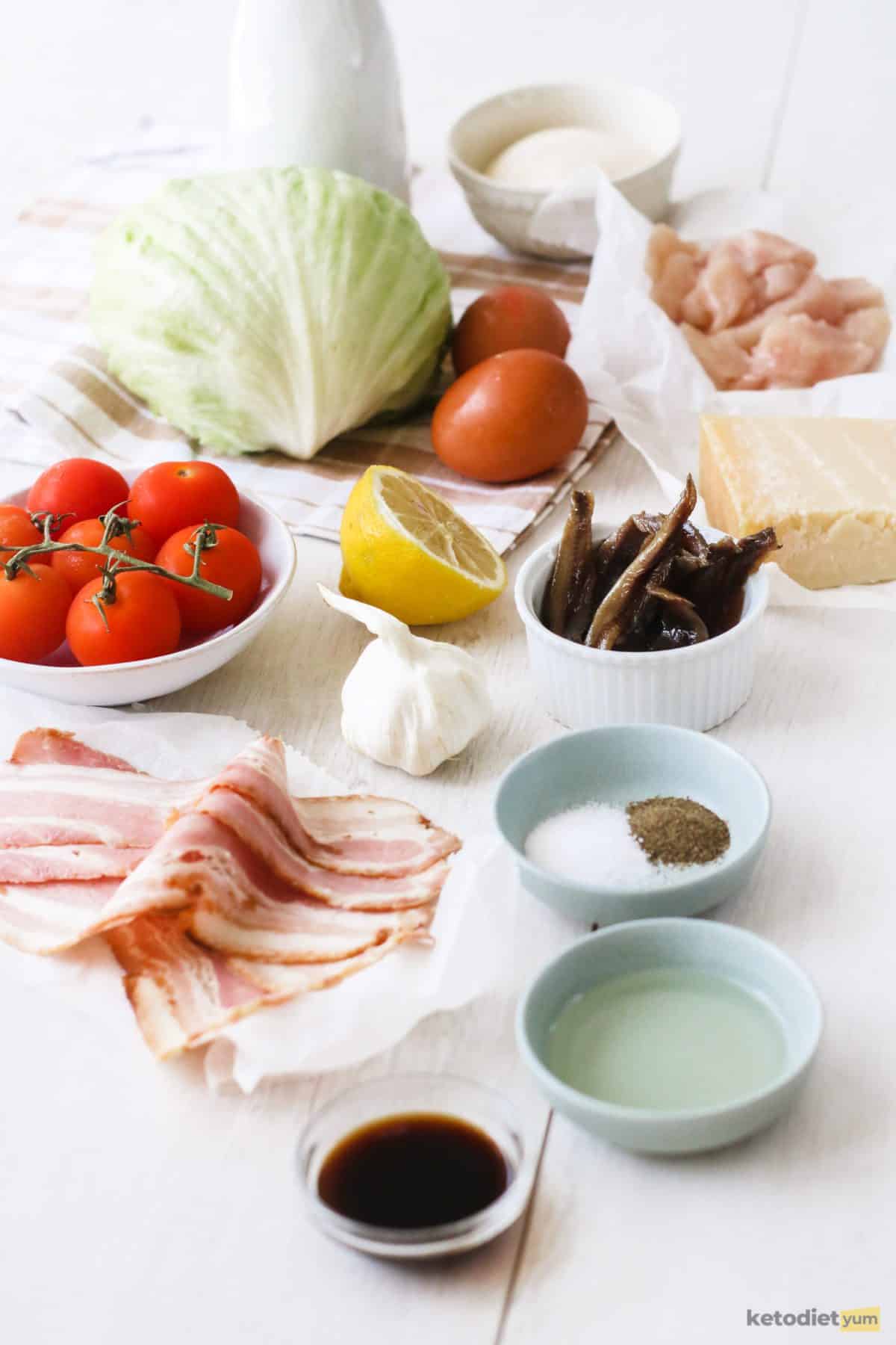 Ingredients arranged on a table to make a Caesar Wedge Salad including bacon, chicken, cherry tomatoes, iceberg lettuce, anchovies, lemon and parmesan cheese.