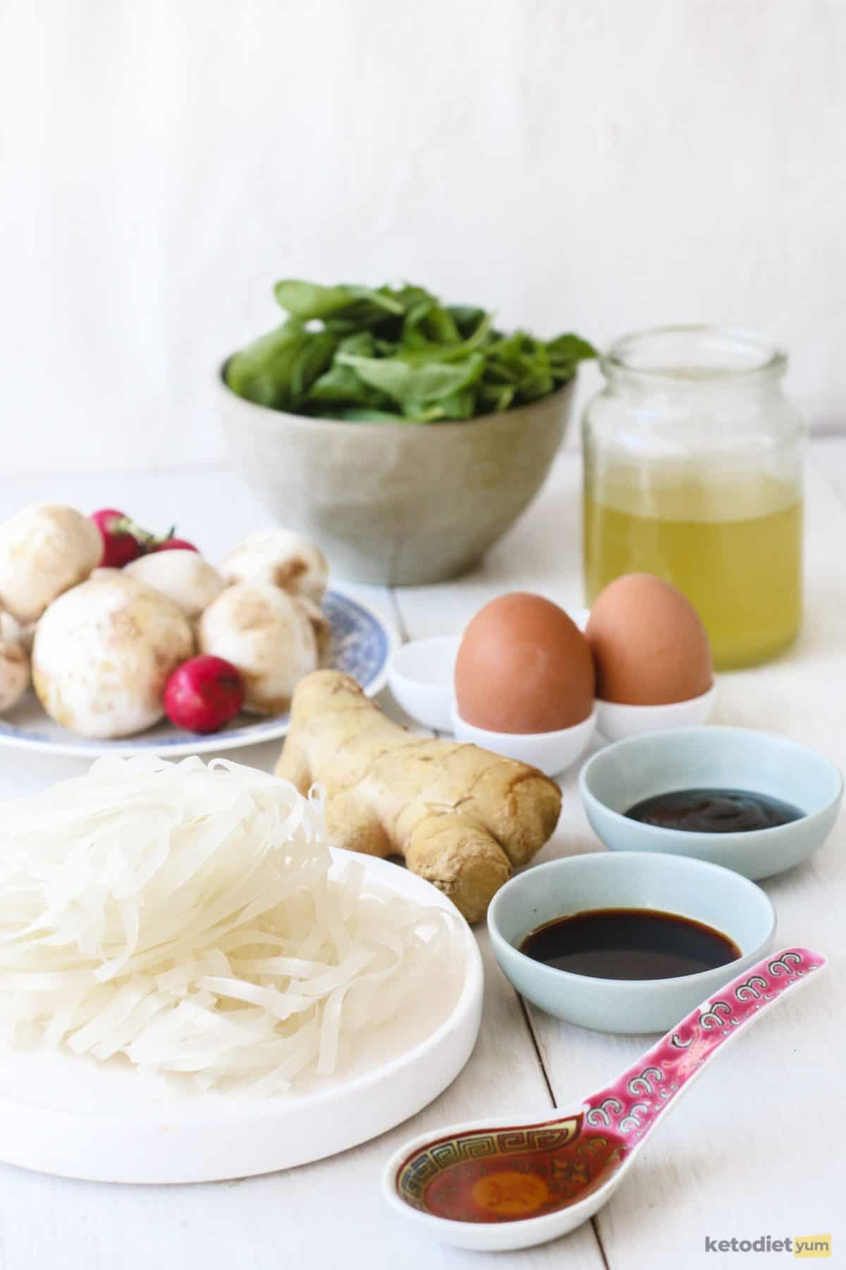 Ingredients arranged on a table to make keto ramen including shirataki noodles, spinach, button mushrooms, eggs, chicken broth, soy sauce and sesame oil