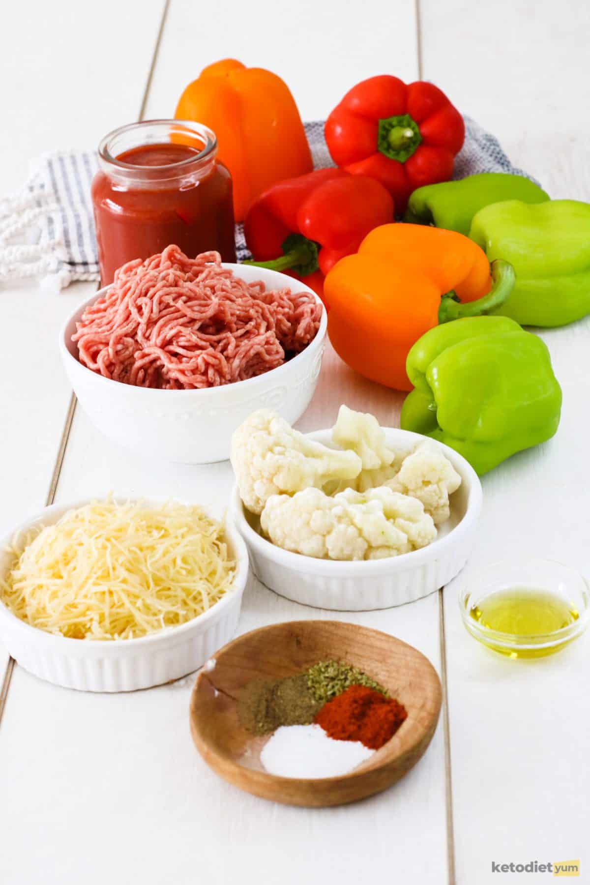 Fresh ingredients arranged on a table including bell peppers, ground beef, cauliflower, cheese, crushed tomatoes, olive oil, herbs and seasonings