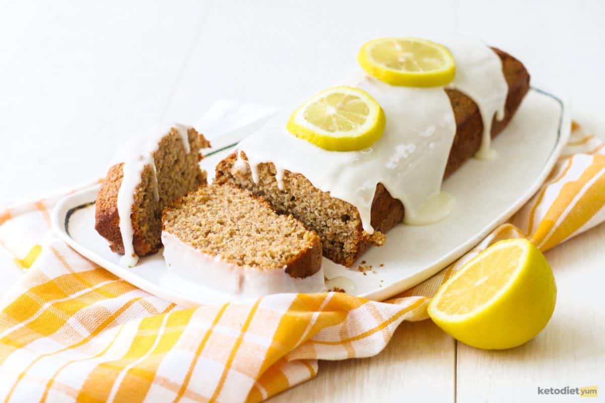 Keto lemon pound cake sliced and served on a white serving plate topped with a white frosting and sliced lemon