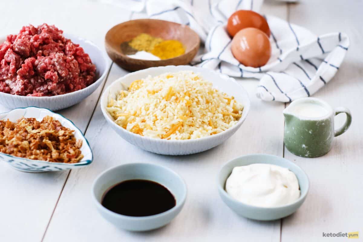 Ingredients arranged on a table used to make keto cheeseburger pies including ground beef, mixed cheese, heavy cream, eggs, mayonnaise, Worcestershire sauce, olive oil, garlic powder, dried onion and ground mustard