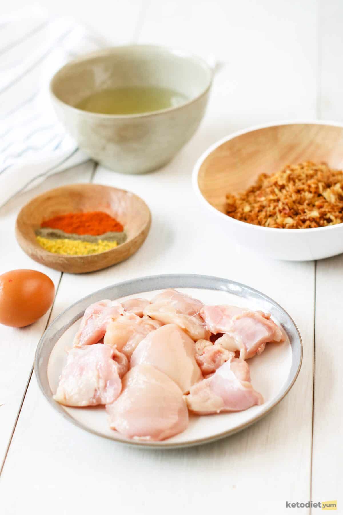 Ingredients arranged on a table to make keto chicken nuggets including chicken, egg, pork rinds, garlic powder, smoked paprika, salt and black pepper