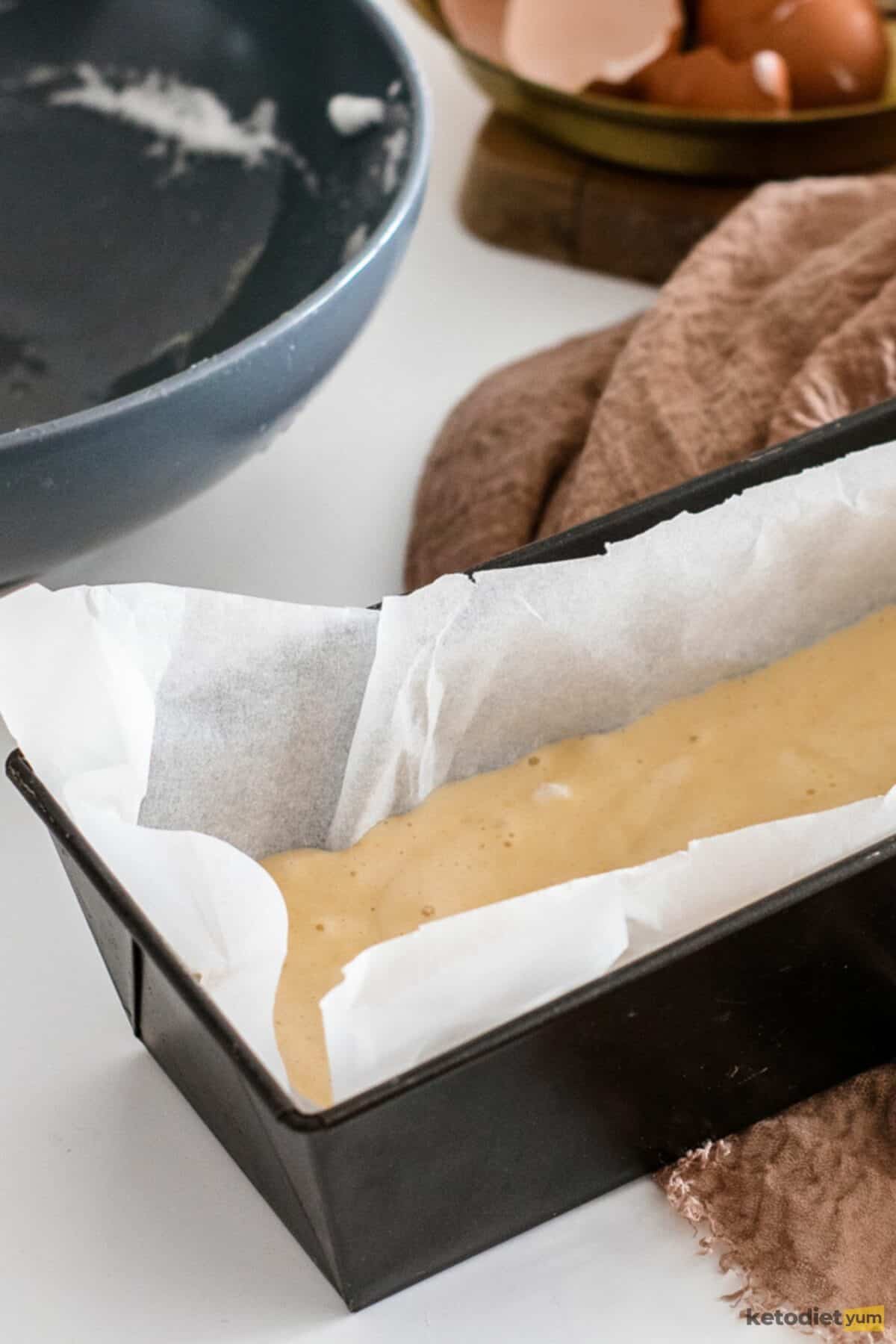 Lined loaf pan filled with batter with empty bowls and egg shells in the background