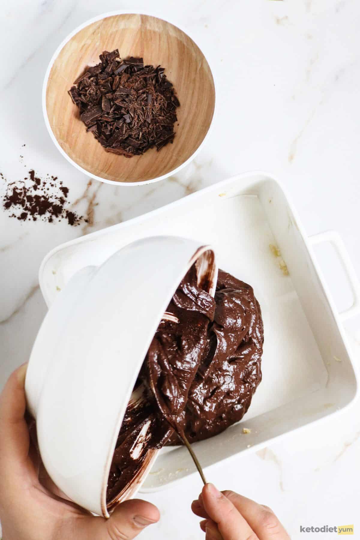 Pouring brownie batter from a bowl into a baking pan with a small bowl of chopped chocolate pieces in the background