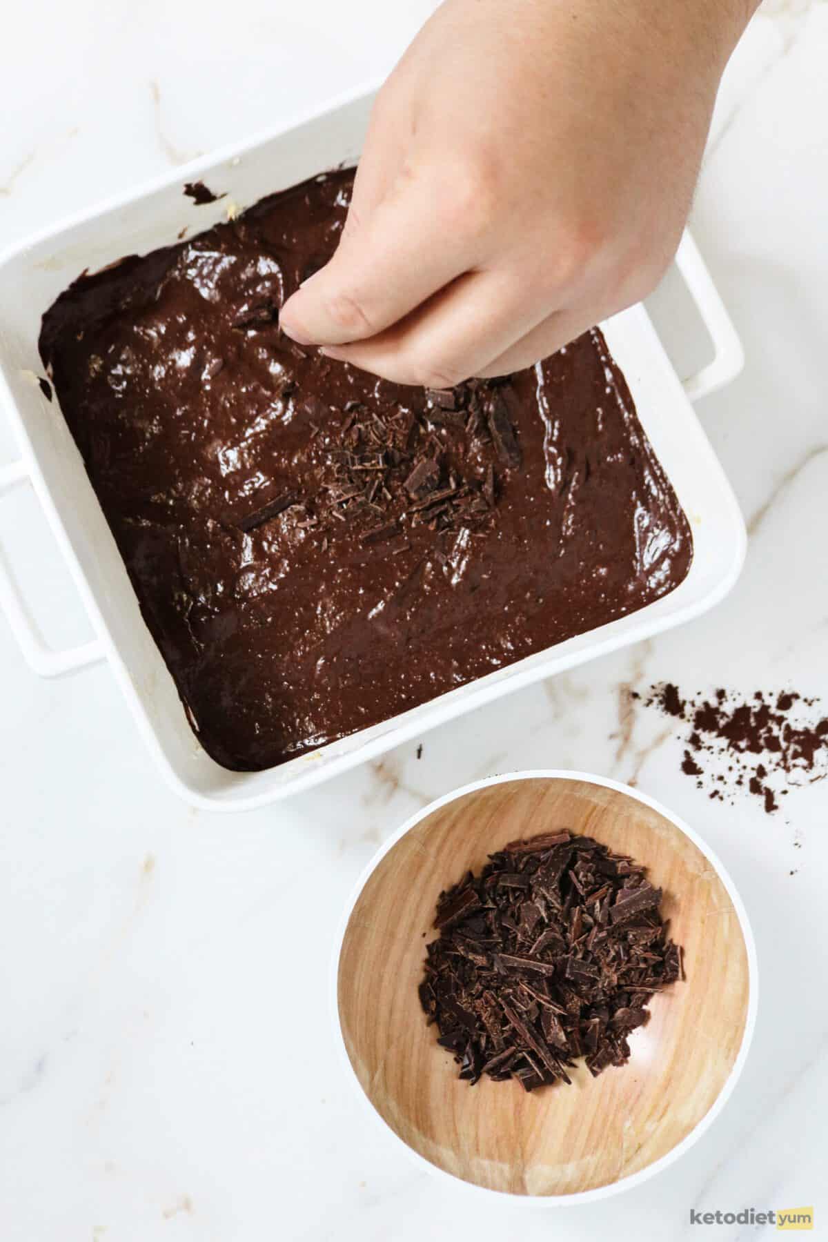Baking pan with brownie batter with a hand sprinkling chocolate over the top and a small bowl of chocolate on a table