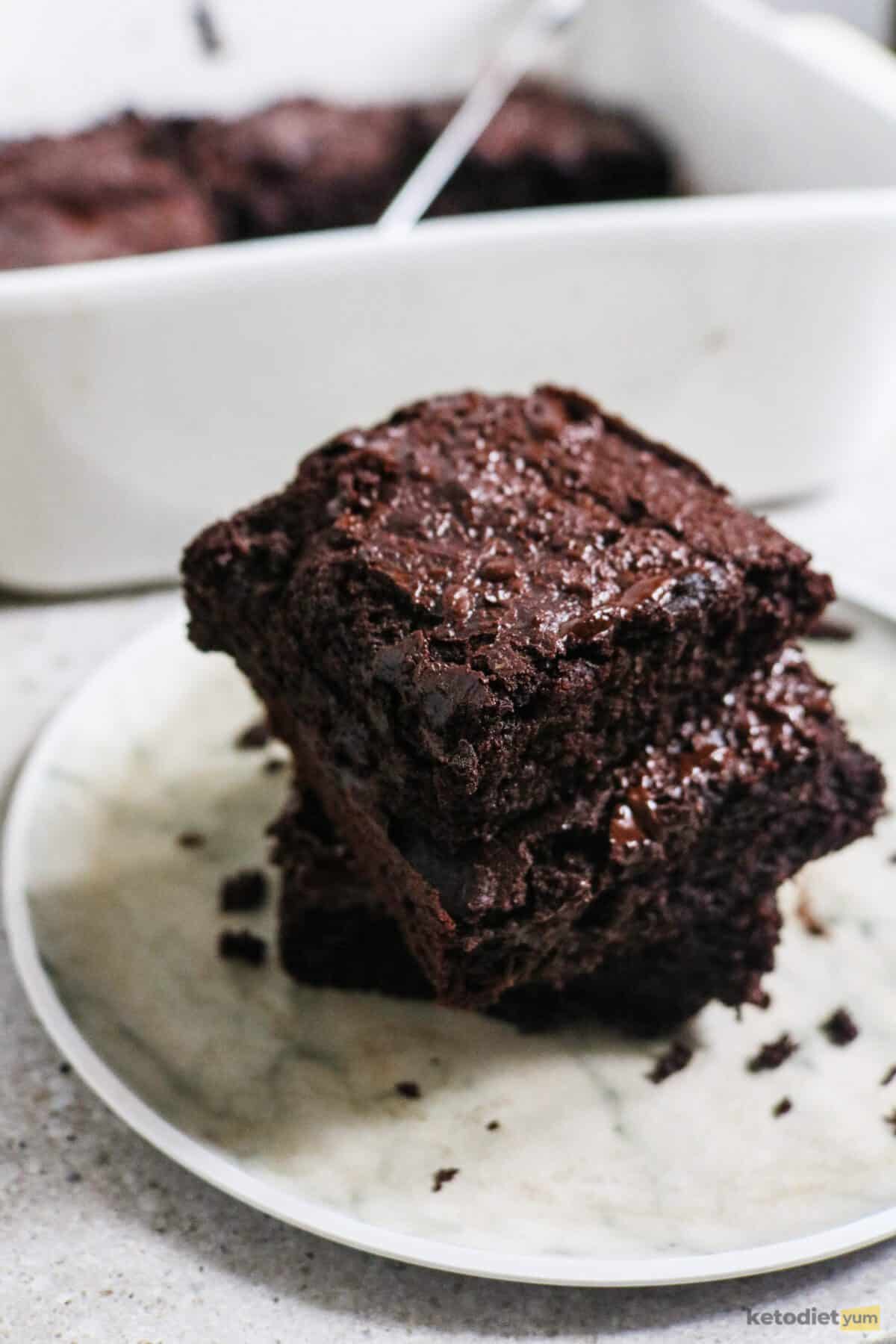 Keto Avocado Brownies Recipe White plate with 3 squares of avocado brownie stacked on top of each other and a baking pan in the background