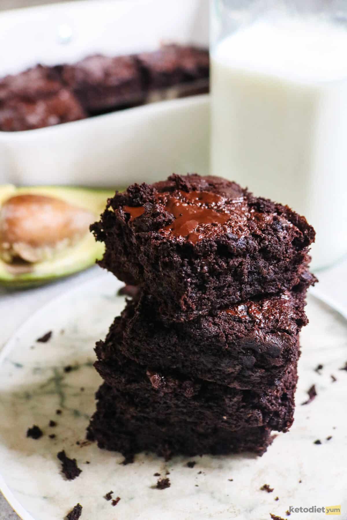 Stack of 3 avocado brownies on a plate with half an avocado, a glass of milk and a baking pan with more brownie in the background