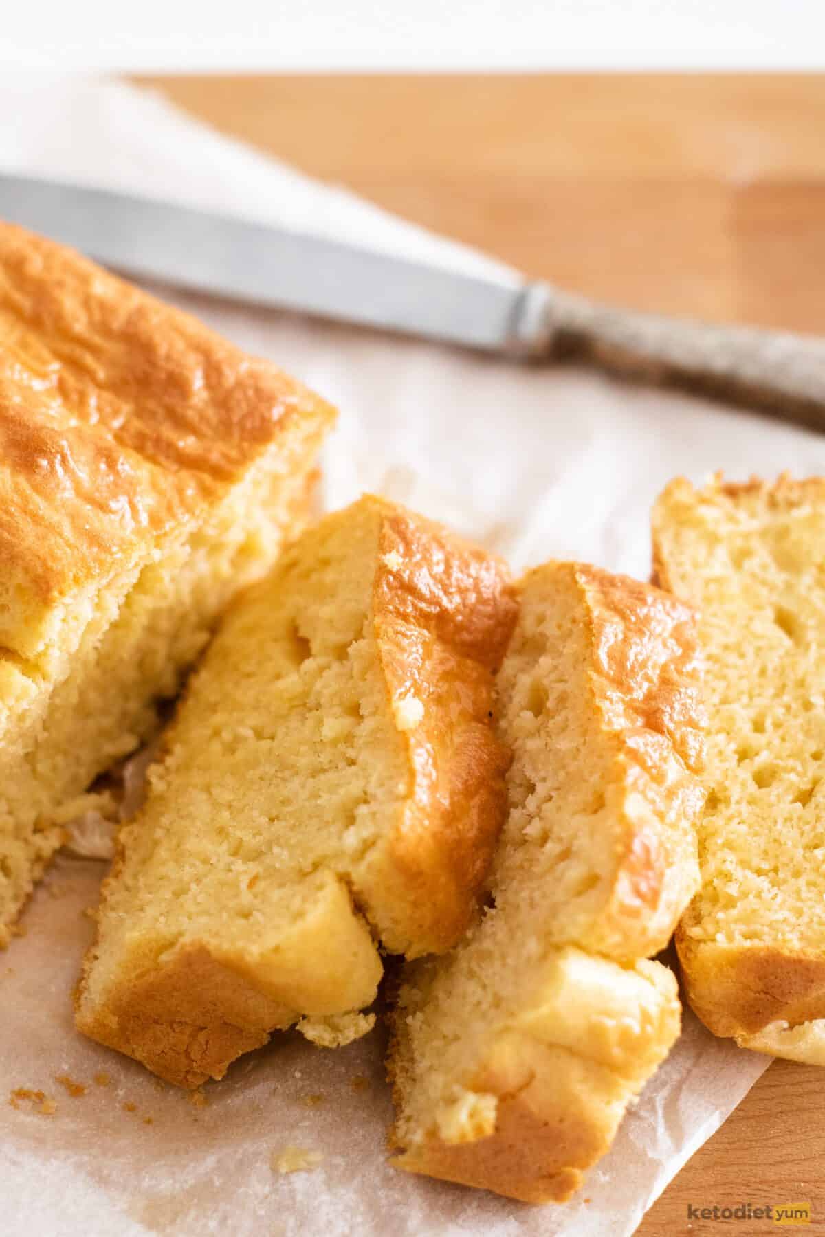 A sliced loaf of bread on top of baking paper sitting on a wooden board next to a butter knife