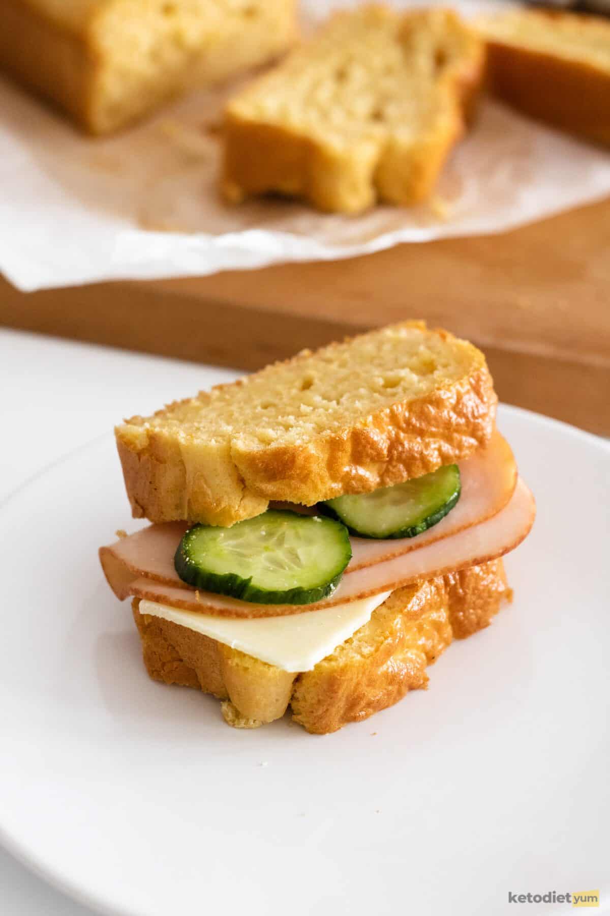 Sandwich with cucumber, ham and cheese on a white plate with a loaf of bread on baking paper in the background