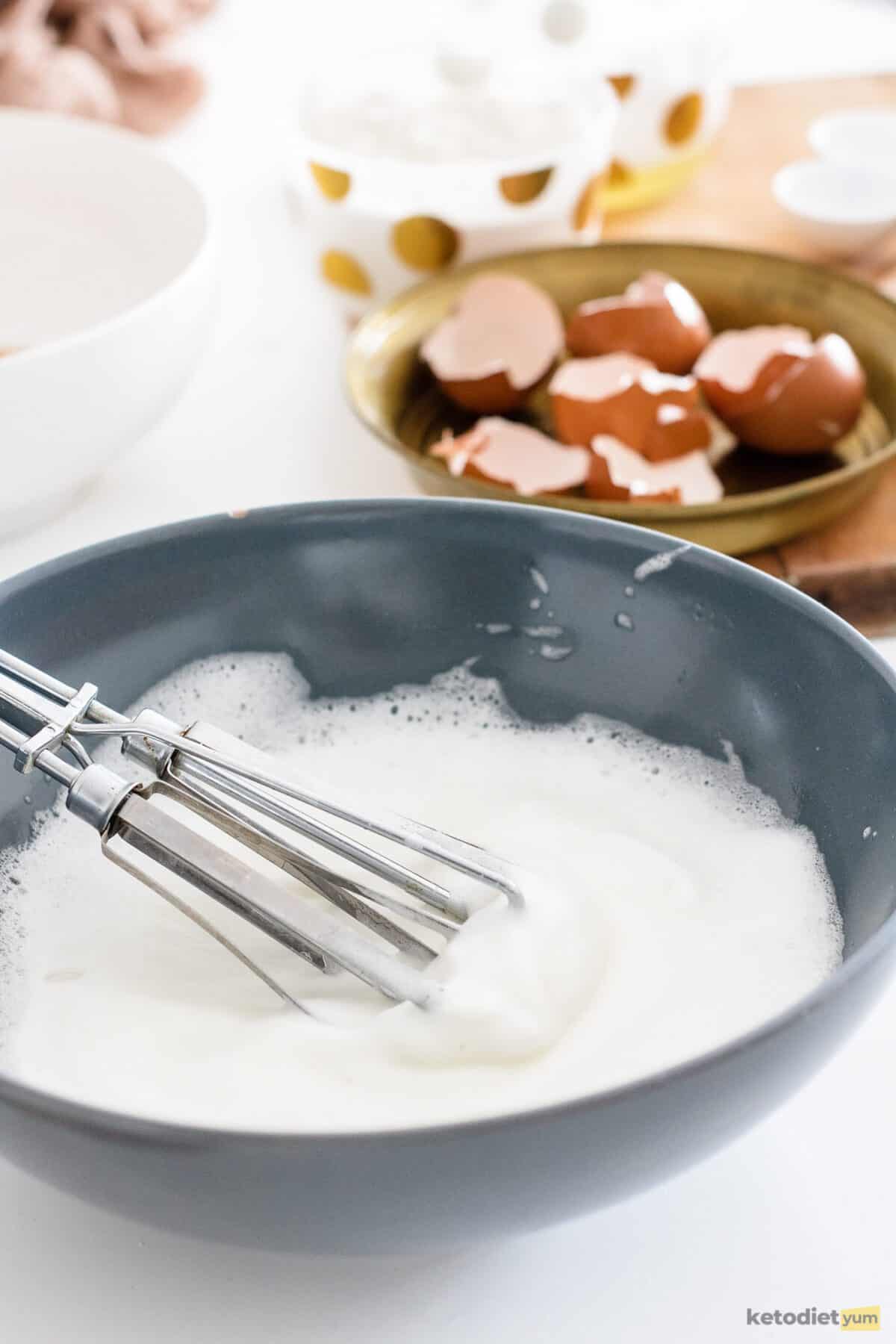 Bowl with egg whites and a whisk with cracked egg shells in the background