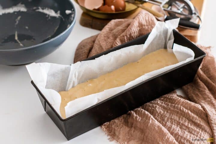 Lined loaf pan filled with batter with empty bowls and egg shells in the background