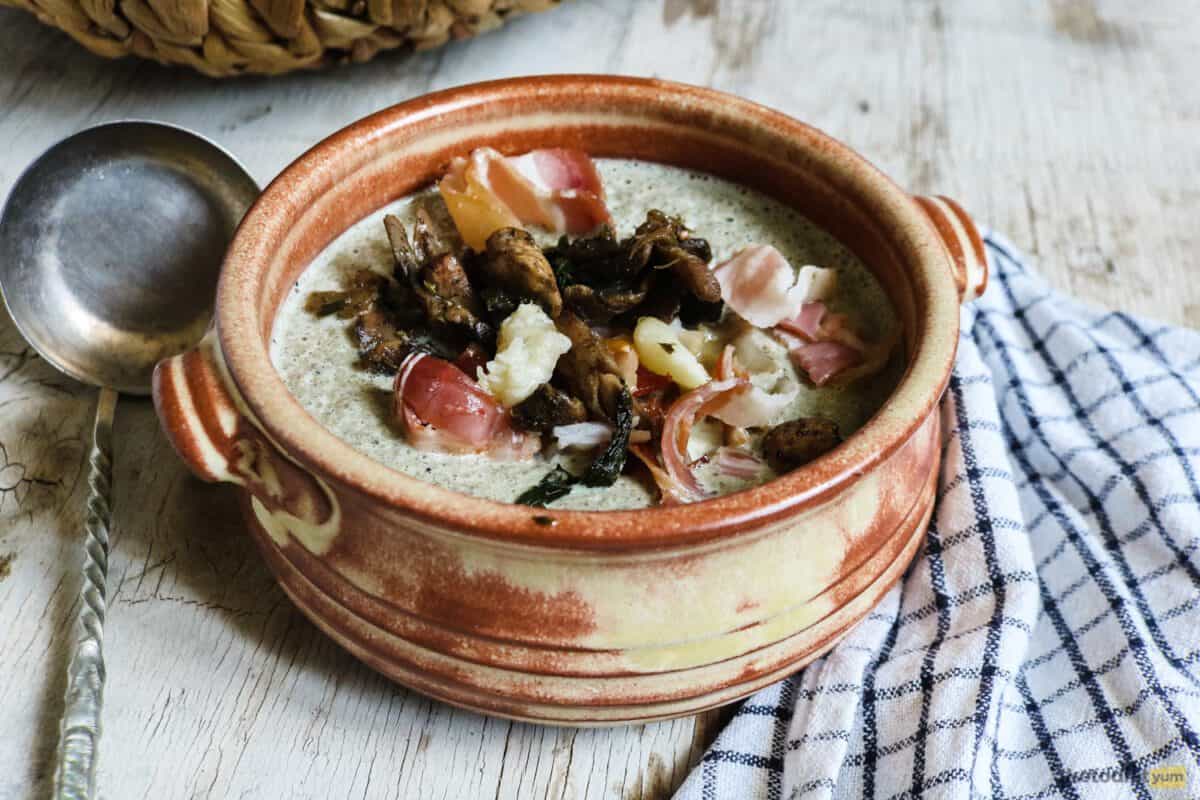 Bowl of keto mushroom soup garnished with bacon and sautéed vegetables sitting on a kitchen table with a ladle and kitchen towel 