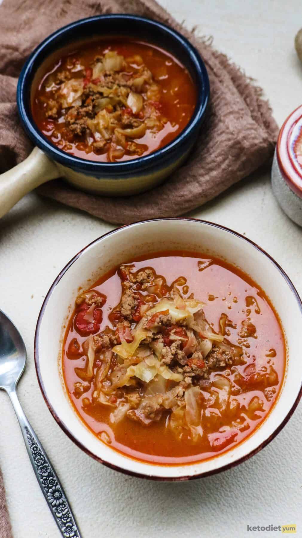 Bowl of tomato cabbage soup on a table with a spoon next to it and a pot of tomato cabbage soup on a kitchen towel in the background