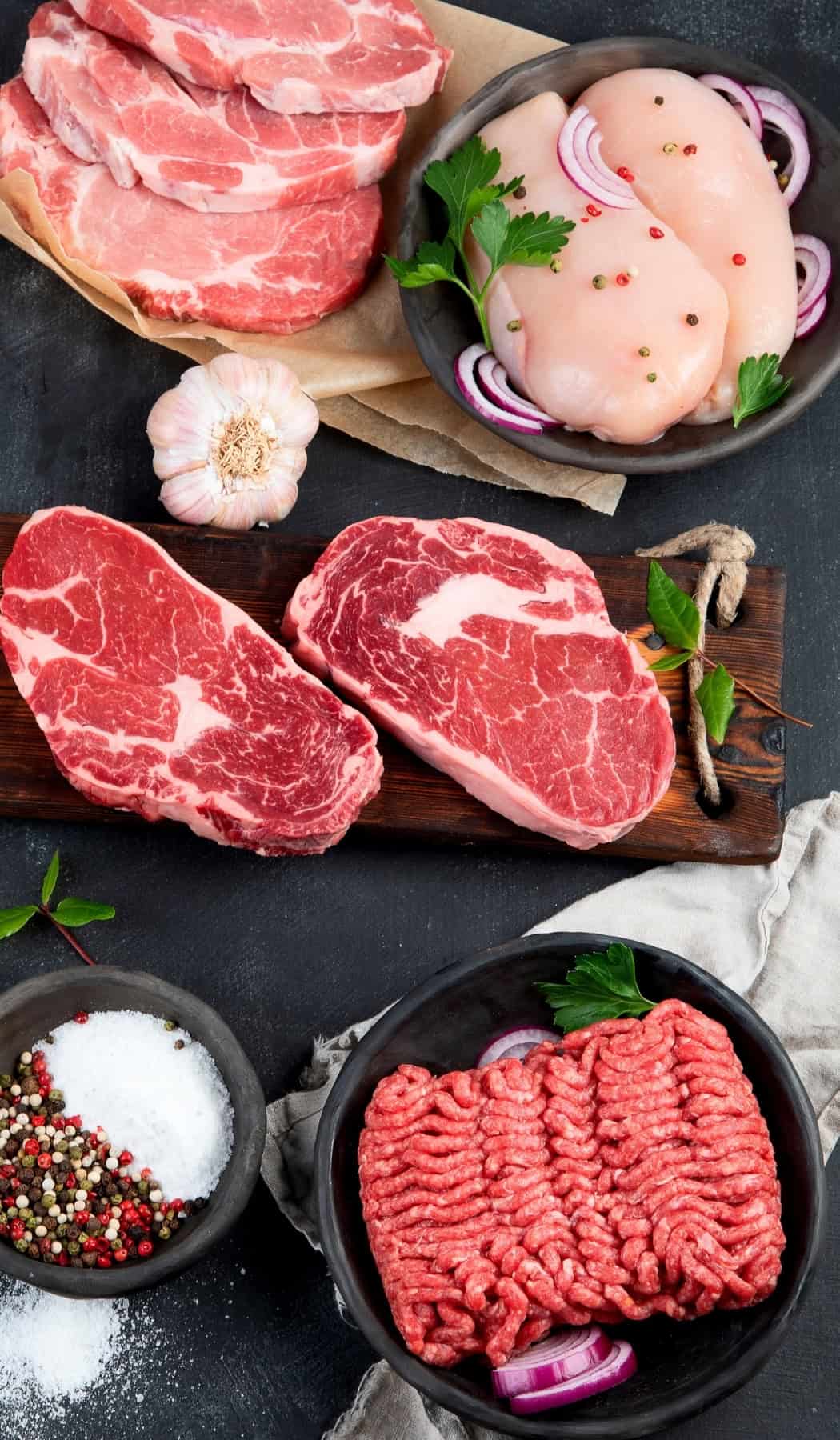 Carnivore Diet for Beginners: Easy Guide & 21-Day Meal Plan Raw cuts of beef, pork, chicken, and ground meat arranged on a dark surface with garlic, salt, pepper, and fresh herbs to show the simple animal foods eaten on the carnivore diet for beginners.