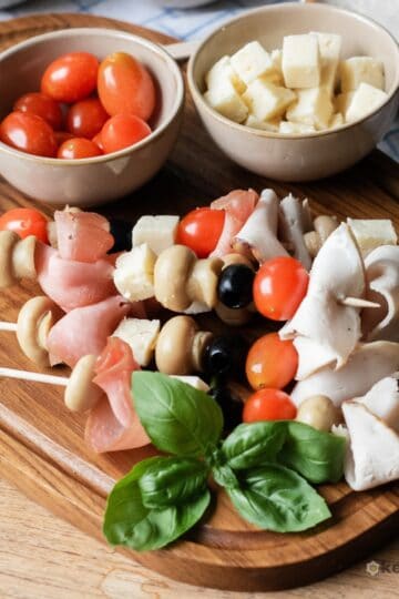 Easy Antipasto Skewers (Low Carb Appetizer) Finished antipasto skewers appetizer on a wooden board, garnished with fresh basil and served with small bowls of cherry tomatoes and cheese cubes.