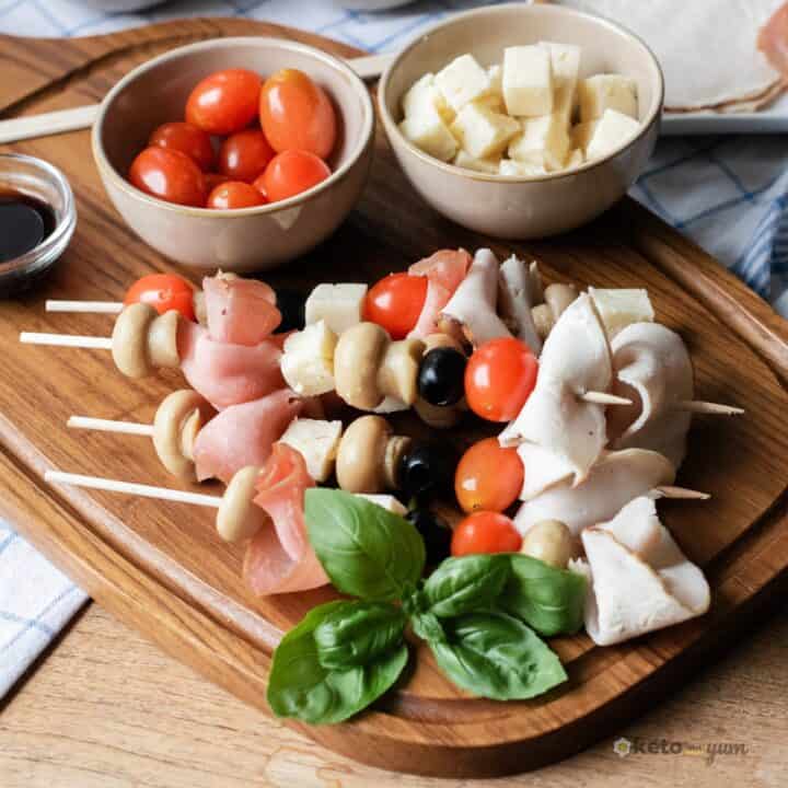 Finished antipasto skewers appetizer on a wooden board, garnished with fresh basil and served with small bowls of cherry tomatoes and cheese cubes.