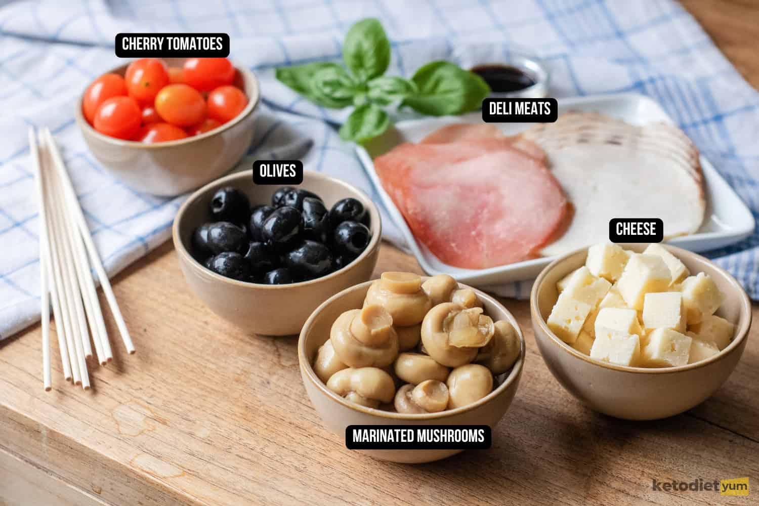 Easy Antipasto Skewers (Low Carb Appetizer) Labeled ingredients for antipasto skewers laid out on a wooden table, including cherry tomatoes, olives, marinated mushrooms, cheese cubes, and deli meats.