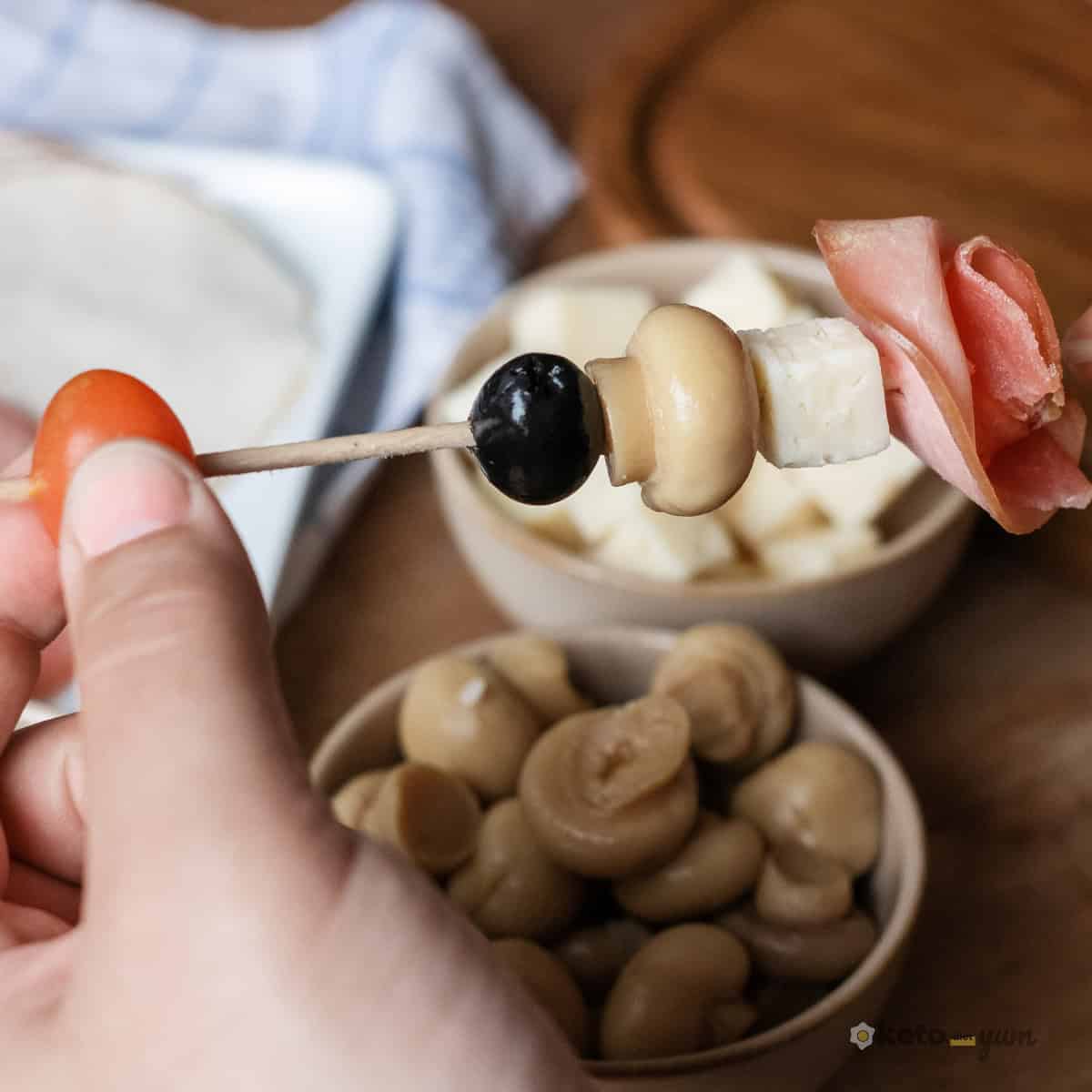 Easy Antipasto Skewers (Low Carb Appetizer) Close-up of a hand assembling an antipasto skewer with a cherry tomato, olive, marinated mushroom, cheese cube, and folded deli meat slice.