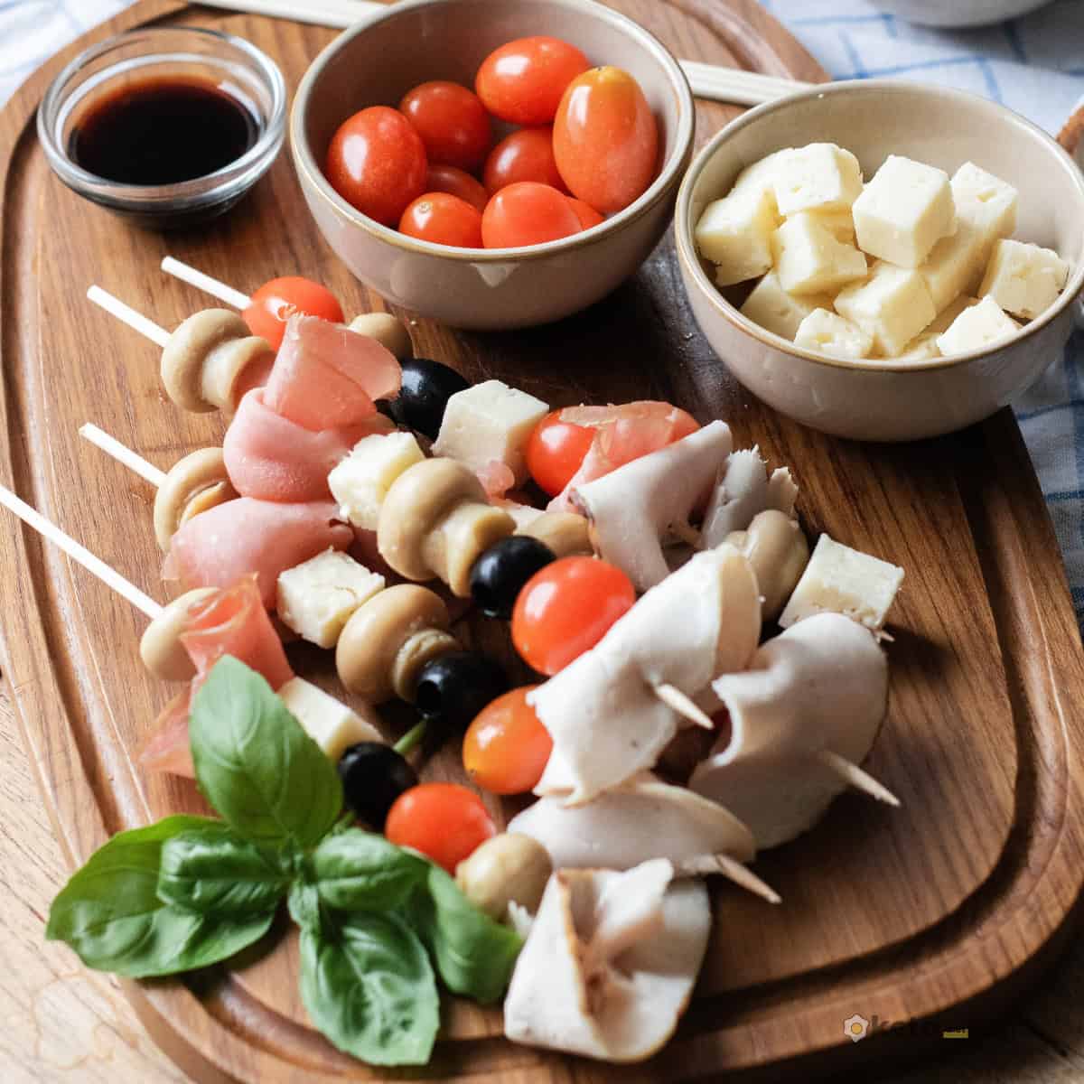 Easy Antipasto Skewers (Low Carb Appetizer) Antipasto skewers appetizer on a serving board surrounded by small bowls of cherry tomatoes, cheese cubes, and balsamic glaze, ready to serve.