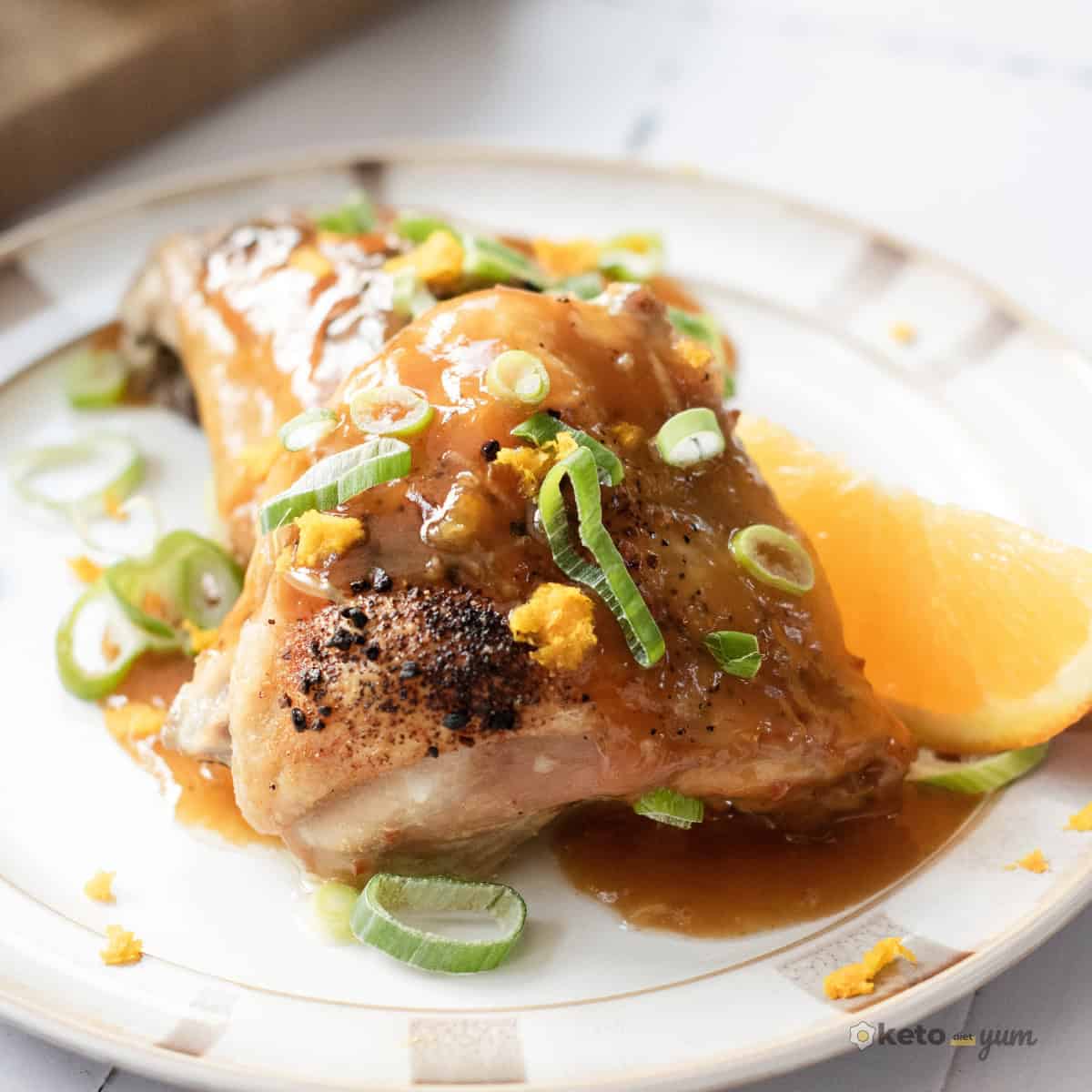 Orange Chicken Thighs (Better Than Takeout) Close-up of crispy orange chicken thigh topped with glossy orange glaze, fresh green onions, and orange zest on a white plate.