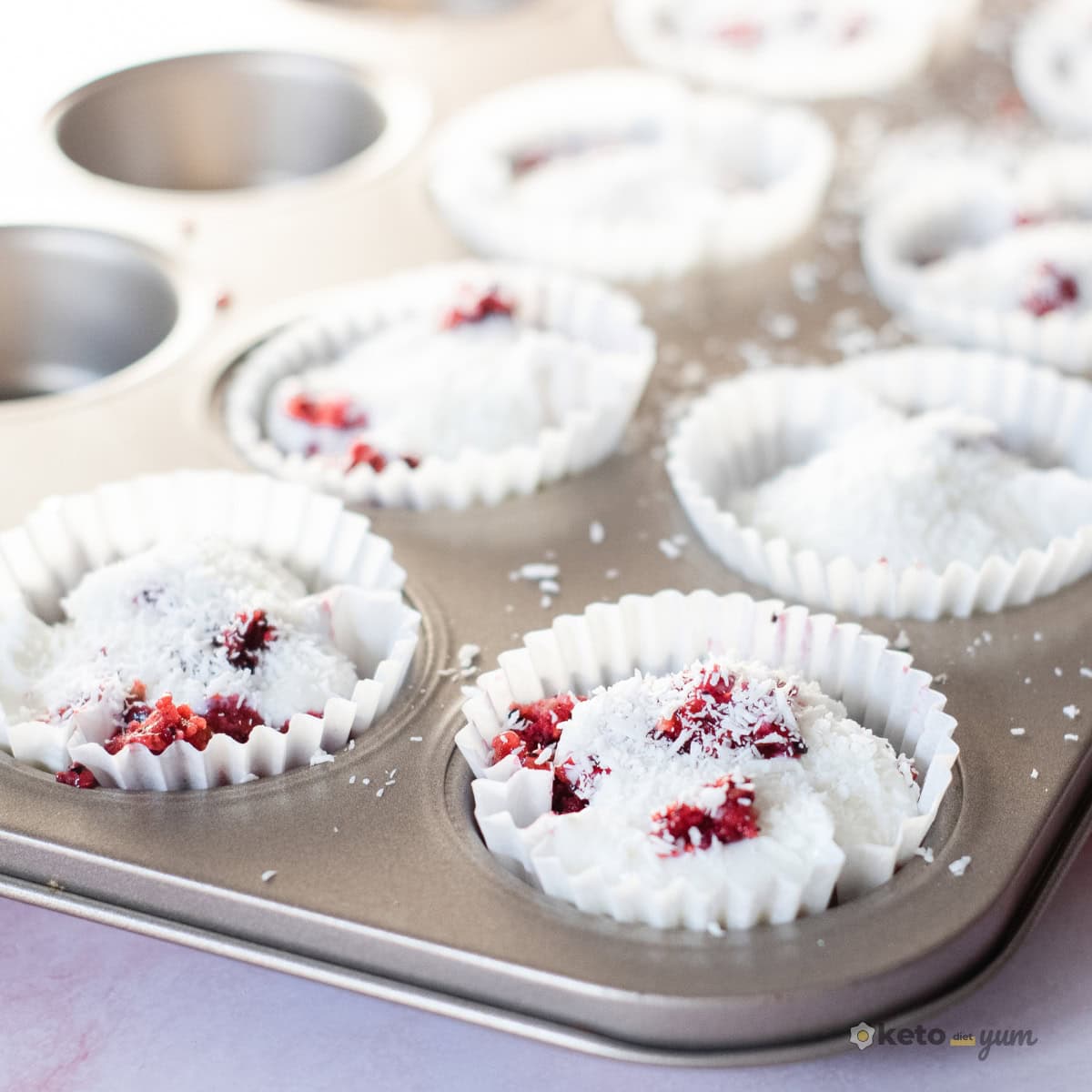 3 Ingredient Frozen Yogurt Bites Muffin tin lined with paper cups filled with coconut-flavored Greek yogurt and berry puree, sprinkled with shredded coconut before freezing.